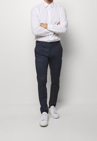 Biała koszula z kołnierzykiem, zapinana na guziki, w połączeniu z ciemnoniebieskimi spodniami w wzory oraz białymi trampkami. Spodnie mają gładką fakturę i prosty krój.