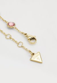 Gouden kettingaccessoire met een roze edelsteen, een kreeftsluiting en een driehoekige hanger met het woord "GUESS" gegraveerd. Duidelijk ontwerp en glanzende afwerking.