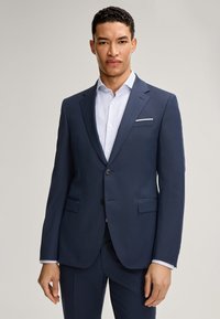 Completo blu scuro con vestibilità su misura, dotato di due bottoni, rever a punta e tasche laterali. Abbinato a una camicia bianca impeccabile.