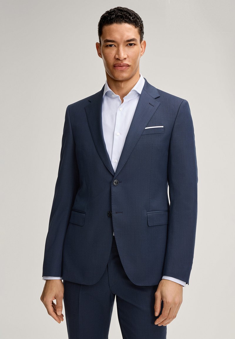 Completo blu scuro con vestibilità su misura, dotato di due bottoni, rever a punta e tasche laterali. Abbinato a una camicia bianca impeccabile.