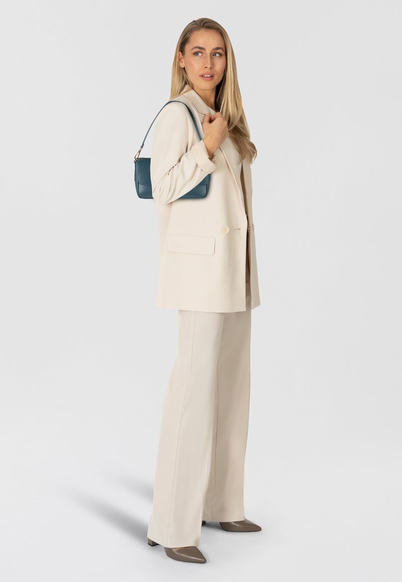 Completo giacca e pantaloni beige, abbinato a una borsa turchese. Tessuto liscio, vestibilità sartoriale, con tasche frontali. Tacchi marroni a punta completano il look.