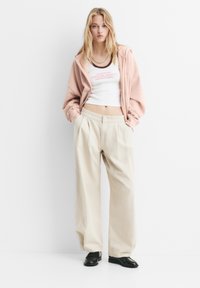 Lichtroze zip-up hoodie over een witte crop top met tekst, gecombineerd met beige wijde broek en zwarte schoenen. Eenvoudige, casual outfit.