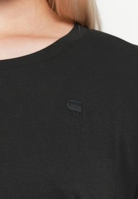 Detailní záběr na osobu v černém tričku s malým, diskrétním vyšívaným logem na hrudi poblíž klíční kosti.