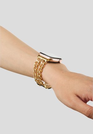 Smartklokke med gullfarget dobbeltdel metallbånd som bæres på venstre håndledd mot en ensfarget grå bakgrunn.