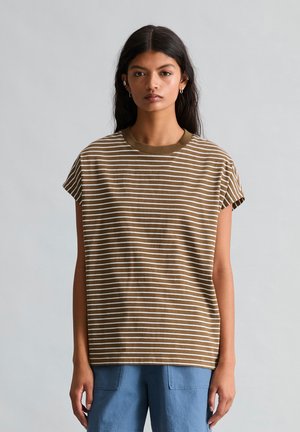 Vrouw met lang donker haar die een bruin-wit gestreepte mouwloze top en een blauwe broek draagt, staand tegen een effen grijze achtergrond.