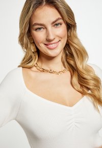 Top bianco a costine con scollatura a V e texture arricciata; una collana di catena dorata e orecchini dorati testurizzati aggiungono accenti metallici.