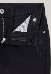 Vaqueros de mezclilla negra con la cremallera parcialmente desabrochada, cinturilla abrochada y forro de los bolsillos que muestra una tela impresa en azul y blanco.