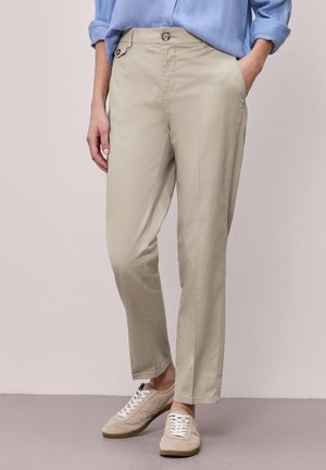 Persoon die beige broek met rechte pijpen draagt, voorzien van een voorste knoop en zijflapzak, een blauwe opgerolde lange mouwen overhemd en beige veter sneakers.