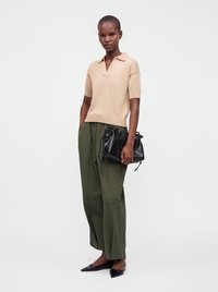 Femme debout portant un haut beige à manches courtes, un pantalon large vert olive, des ballerines noires pointues, tenant un sac à main noir.