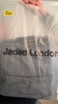 Przezroczyste plastikowe opakowanie zawiera złożoną odzież z ciemnymi poziomymi paskami, na której wyraźnie widoczny jest napis "Jaded London".