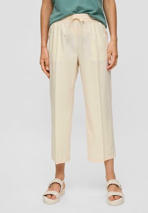 Nainen pukeutuneena beigenvärisiin lyhyisiin housuihin, joissa on joustava vyötärönauha ja kiristysnauha, yhdistettynä kermanvärisiin punottuihin sandaaleihin ja vihreään yläosaan.