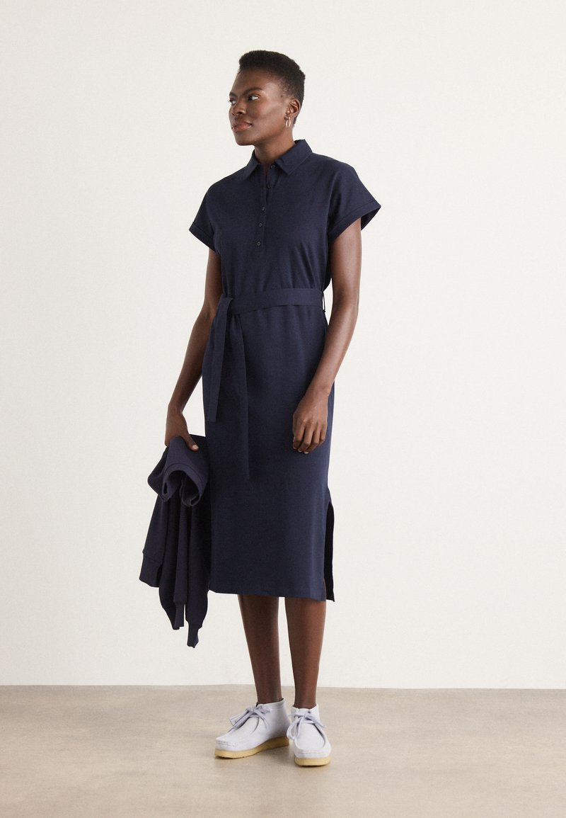 Granatowa bawełniana sukienka polo z krótkimi rękawami, przewiązana w talii, z bocznymi rozcięciami, w połączeniu z jasnoszarymi butami i dopasowanym swetrem.
