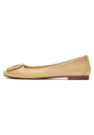 Chaussure plate en cuir beige avec un petit talon et une grande boucle décorative sur l'avant arrondi, montrée de profil sur fond blanc.