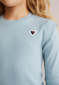Chemise à manches longues côtelée bleu clair avec un petit motif de cœur brodé en bleu marine, marron et blanc sur la poitrine.