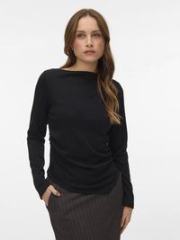 Žena s dlouhými hnědými vlasy nosí černý dlouhý rukáv a hnědou sukni s proužky, stojí s jednou rukou v kapse před bílým pozadím.