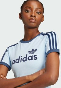 Vaaleansininen puuvillapaitapusero, jossa tummansiniset yksityiskohdat, pyöreä kaula-aukko ja kolme pystysuoraa raitaa hartioilla. Puserossa on tumma "adidas"-logo.