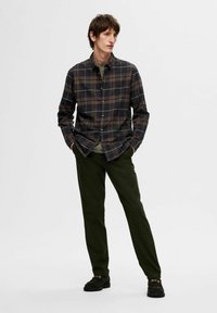 Camicia a quadri in tonalità scure con righe bianche e marroni, abbinata a pantaloni verde oliva. Il modello indossa scarpe nere e si trova con le mani in tasca.