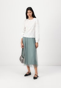 Blusa bianca ricamata a fiori con maniche a sbuffo, abbinata a una gonna plissettata color teal. Sandali neri in stile Birkenstock con dettagli e una borsa argentata.