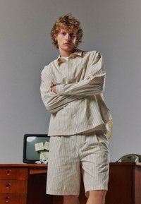 Jeune personne aux cheveux bouclés, les bras croisés, portant une chemise rayée crème et un short assorti dans un bureau avec une télévision vintage et un téléphone.