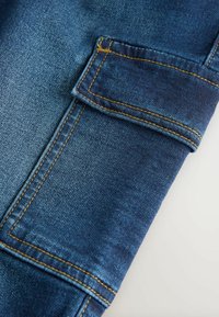 Tumedat sinelid kanga, mis on tume sinine, koos klapiga taskuga, kontrastsete kollaste õmblustega ja siledaks tekstuuriga. Omab kohandatud lõiget.