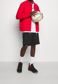 Rode lichtgewicht jas met knoopsluiting, witte T-shirt, zwarte shorts, witte sokken en zwarte sneakers met rode accenten, terwijl hij een basketbal vasthoudt.