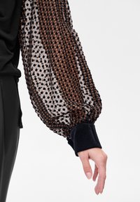 Bluzka z bufkami wykonana z przezroczystej tkaniny w geometryczny wzór w kolorach brązowym i czarnym, z czarnym, aksamitnym mankietem i dopasowanym krojem.