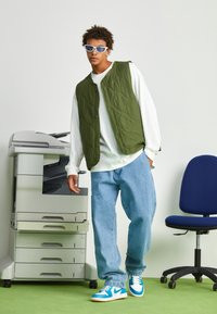 Green quilted vest over a white long-sleeve shirt, light blue jeans, and blue Nike sneakers. Soft textures, modern design with buttons.