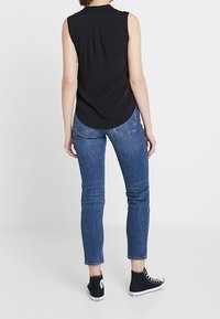 Haut noir sans manches avec des plis au dos, associé à un jean bleu ajusté et des chaussures montantes noires. La tenue présente un design décontracté.