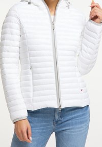 Kobieta ubrana w białą pikowaną kurtkę na zamek z kapturem oraz jasnoniebieskie jeansy, pozująca z jedną ręką przy twarzy, drugą swobodnie opuszczoną.