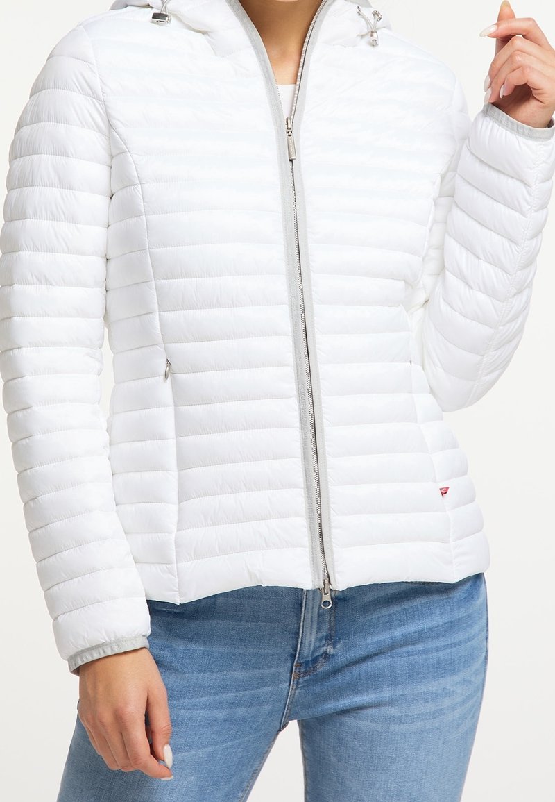 Kobieta ubrana w białą pikowaną kurtkę na zamek z kapturem oraz jasnoniebieskie jeansy, pozująca z jedną ręką przy twarzy, drugą swobodnie opuszczoną.