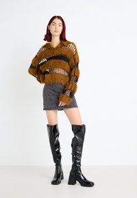 Pull marron duveteux avec des rayures en maille noire, mini-jupe grise avec des détails à clous, et bottes noires à talons montantes avec une finition brillante.