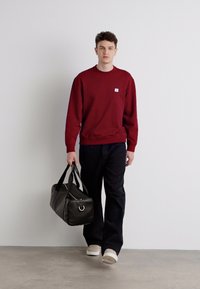 Felpa bordeaux, pantaloni neri a gamba larga, sneakers crema e borsa nera a tracolla con dettagli in argento, in piedi su uno sfondo neutro.