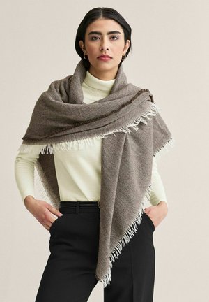 Nainen, jolla on päällään kermaan vivahtava pooloneule, mustat housut ja suuret ruskeat hapsulliset viitat hartioillaan, beige tausta.