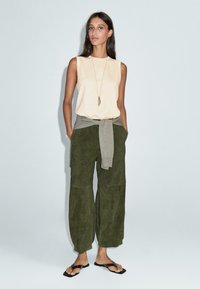 Una donna indossa un top beige senza maniche, pantaloni larghi oliva, un maglione grigio legato in vita, sandali neri e una lunga collana con pendente.