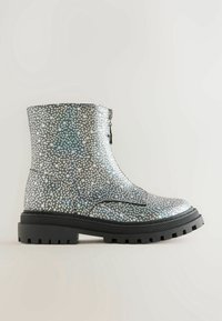 Botte argentée montant à la cheville avec petits motifs d'étoiles blanches, semelle épaisse noire et fermeture éclair avant sur fond uni.
