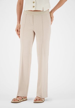 Beige zigzagpatroon broek gedragen met beige sandalen en een mouwloos topje met knopen, staand tegen een effen witte achtergrond.