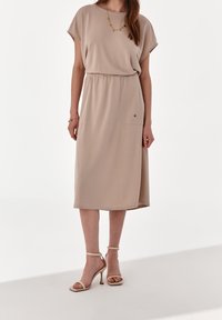 Béžové midi šaty s krátkým rukávem, elastickým pasem a dvěma předními kapsami. Stylizované s nahými lodičkami na vysokém podpatku a jemným náhrdelníkem.