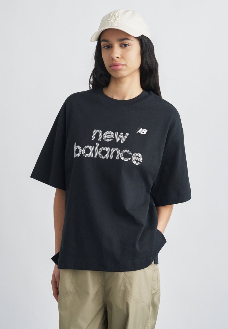 Νεαρή γυναίκα που φοράει μαύρο t-shirt New Balance, μπεζ παντελόνι και λευκό καπέλο μπέιζμπολ, στέκεται με το ένα χέρι στην τσέπη μπροστά σε απλό φόντο.