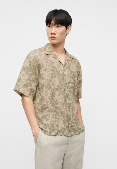 Νεαρός άνδρας με κοντά μαύρα μαλλιά, φοράει μπεζ κοντομάνικο πουκάμισο με λουλούδια και ανοιχτό μπεζ παντελόνι με τα χέρια στις τσέπες, κοιτάζοντας πλάγια.