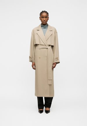 Femme portant un trench-coat beige ceinturé sur un col roulé bleu clair, un pantalon noir et des chaussures pointues noires, debout devant un fond uni.