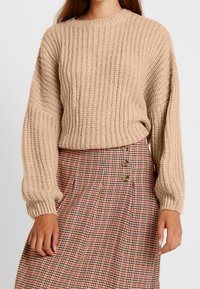 Pull beige en maille avec texture côtelée et manches larges, associé à une jupe à carreaux avec des détails de boutons dans des tons rouges et bruns.