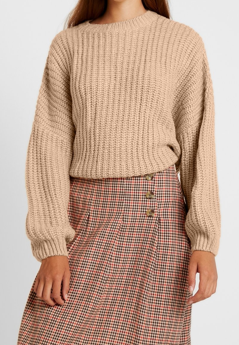 Pull beige en maille avec texture côtelée et manches larges, associé à une jupe à carreaux avec des détails de boutons dans des tons rouges et bruns.