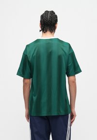 Muškarac s pletenicama, odjeven u tamnozeleno majicu kratkih rukava i tamnoplave hlače s bijelim prugama, stoji okrenut leđima na jednobojnoj pozadini.