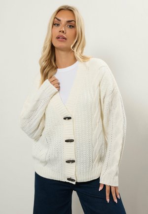 Weißer Strickcardigan mit lässigem Schnitt, Zopfmuster und großen schwarzen Knöpfen auf der Vorderseite. Mit langen, gerippten Ärmeln und offenem Ausschnitt.