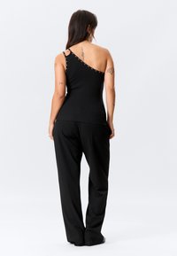 Μαύρο τοπ ενός ώμου με μεταλλικά κουμπιά κατά μήκος του λουριού, συνδυασμένο με χαλαρό μαύρο παντελόνι, που δείχνει μια λεία, εφαρμοστή γραμμή.