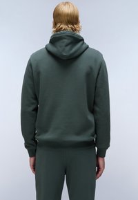 Donkergroen hoodie met een zachte textuur en een aansluitende pasvorm. Kenmerkt zich door een grote, verstelbare capuchon en ribgebreide manchetten en onderkant.