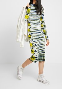 Robe à manches longues teintée par nouage, avec des rayures vertes, jaunes et bleues, fabriquée en tissu doux. Portée avec des baskets blanches et une veste légère.