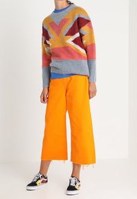 Pull tricoté avec un motif Union Jack en nuances de rouge, jaune et bleu ; associé à un pantalon large de couleur orange vif et des baskets noires.