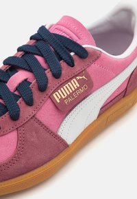 Tenisky PUMA Palermo kombinují semiš a kůži v růžové a bílé barvě, mají námořnické šněrování, gumovou podrážku a zlatý detail loga.