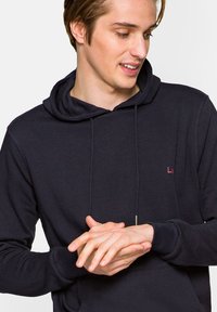 Ciemnoniebieska bluza z kapturem, wykonana z miękkiego materiału, z przednią kieszenią i regulowanymi sznurkami. Posiada małe logo na klatce piersiowej.